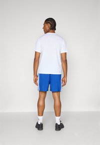 Tricou alb cu mânecă scurtă și guler rotund, asortat cu pantaloni scurți albaștri cu dungi laterale, purtat cu șosete albe și pantofi sport negri.
