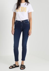 Vit t-shirt med "LÖRDAG" i gul text, kombinerad med mörkblå skinny jeans och svarta sandaler med silverfärgade spännen.