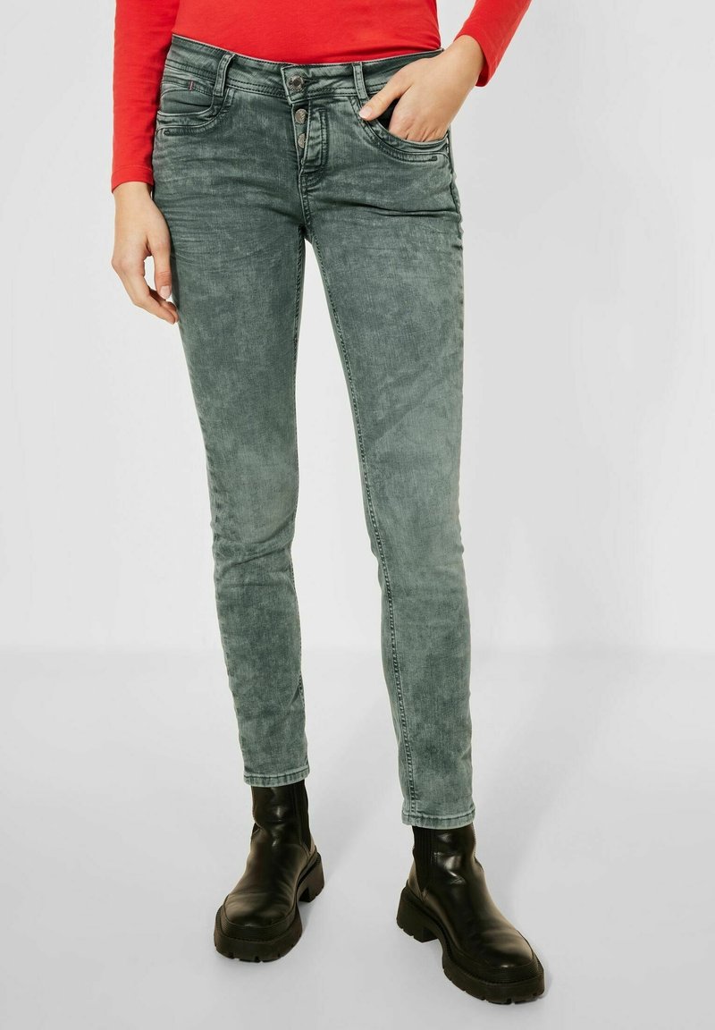 Jean skinny en denim vert délavé, avec deux poches avant et une fermeture à boutons. Associé à des bottines noires.