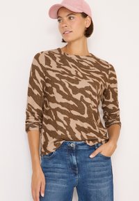 Braun und beige langärmliges Oberteil mit einem Tigerstreifenmuster, kombiniert mit blauen Jeans. Das Modell trägt außerdem eine pinkfarbene Mütze.