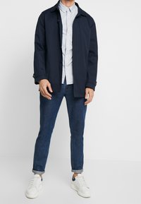 Námořnická lehká bunda s límcem, zapínáním na knoflíky a ohrnutými rukávy, pod kterou je světlomodrá košile, doplněná tmavými džíny a bílými teniskami.