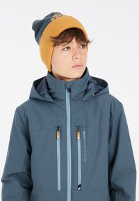 Giacca impermeabile blu con colletto alto, due tasche frontali con zip, tirazip arancioni e un berretto grigio e giallo.