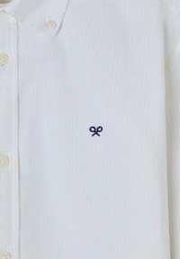 Camisa blanca con botones y textura a rayas verticales, que presenta un pequeño logo de tijeras azules bordado en el lado izquierdo del pecho.