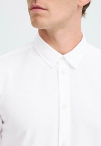 Camisa blanca de botones con cuello estándar, con un tejido texturizado y cinco botones. Vista en primer plano destacando el escote.