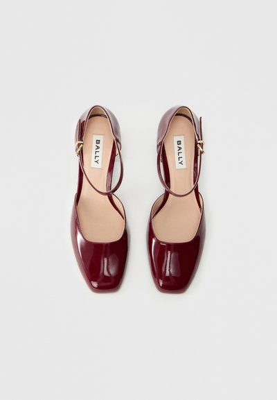 Chaussures en cuir verni bourgogne avec un bout carré, un talon ouvert et une sangle ajustable ornée d'un accent en métal doré. Logo de la marque visible à l'intérieur.