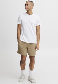 Witte katoenen t-shirt, beige textuurbroekje, lichte sneakers en witte kousen tot aan de kuiten. Zichtbare tatoeage op het rechterbeen.