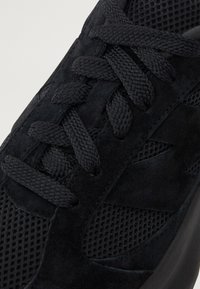 Gros plan d'une basket noire avec des lacets texturés, des empiècements en suède et un tissu mesh respirant sur la partie supérieure de la chaussure.