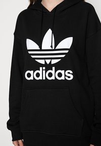 Juoda gobtuvė su didele balta Adidas trefoil logotipu ir tekstu. Pagaminta iš minkštos medžiagos, turi priekinę kišenę ir stringus gobtuve.