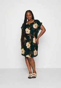 Vestido floral com fundo verde escuro, apresentando padrões de peônias em creme e bege. Mangas curtas, cintura com cordão e comprimento até ao joelho.