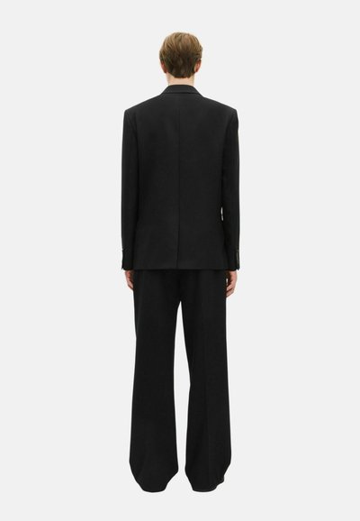 Traje negro a medida que presenta un abrigo largo y pantalones de pierna ancha. El tejido tiene una textura suave con un detalle mínimo.