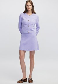 Lila strukturierte Strickjacke mit goldenen Knöpfen und zwei Vorderaschen, kombiniert mit einem passenden A-Linien-Rock und Ballerinas im Leopardenmuster.