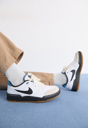 Personne portant des baskets blanches Nike avec le logo noir et un pantalon beige, assise sur un tapis bleu sur fond blanc.