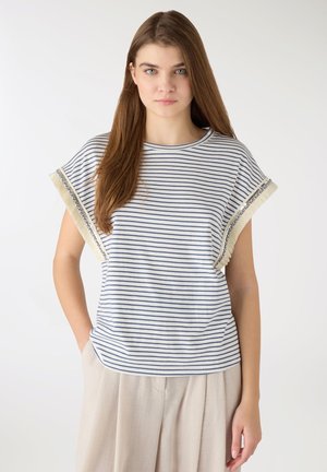 Giovane donna con lunghi capelli castani che indossa una maglietta a maniche corte bianca e blu a righe e pantaloni beige larghi, in piedi davanti a uno sfondo neutro.