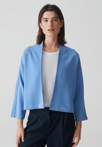 Veste courte en bleu clair, avec une coupe décontractée, des manches larges et une texture lisse. Portée sur un top à côtes blanc et un pantalon foncé.