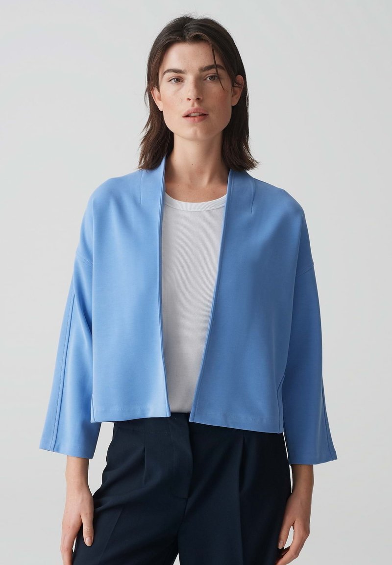 Veste courte en bleu clair, avec une coupe décontractée, des manches larges et une texture lisse. Portée sur un top à côtes blanc et un pantalon foncé.