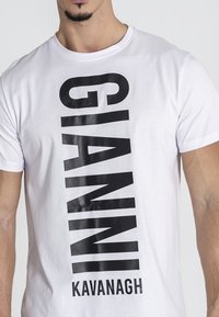 Camiseta blanca de manga corta con un gran diseño gráfico vertical en negro que dice "GIANNI KAVANAGH". Textura de algodón suave.