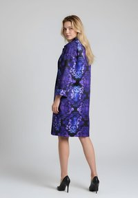 Abrigo morado con estampado y acabado liso y brillante; llega hasta la rodilla, con grandes remolinos estilizados y mangas tres cuartos. Tacones altos negros complementan el conjunto.