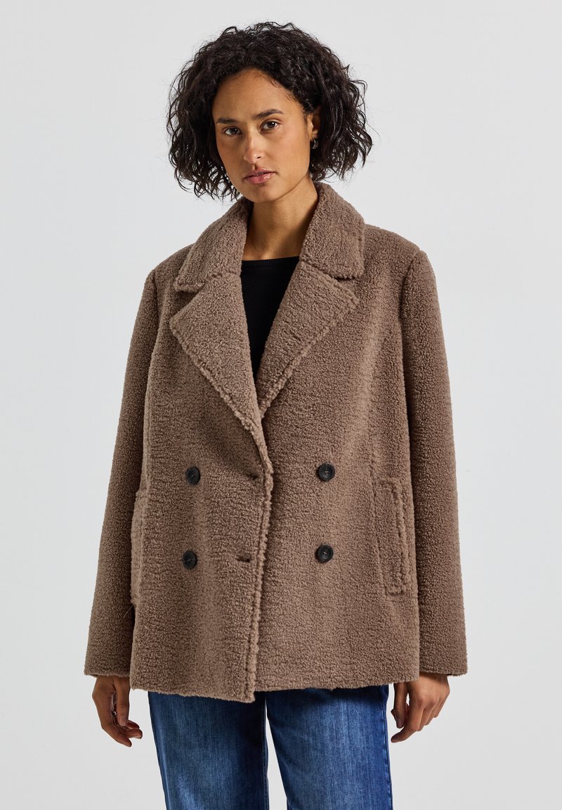 Kvinna iklädd en brun dubbelknäppt teddyrock med svarta knappar, svart topp och blå jeans, stående mot en neutral ljus bakgrund.