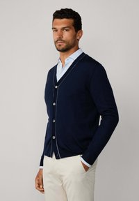 Cardigan blu navy con scollatura a V, chiusura con bottoni e dettagli in bianco. Indossato sopra una camicia a righe azzurra chiara. Maniche lunghe, texture liscia.