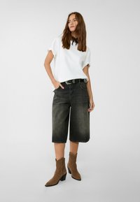 Camicia bianca a maniche corte con orli arricciati, culotte di jeans neri, stivaletti in suede marrone con punta affusolata, cintura nera con dettagli argentati.