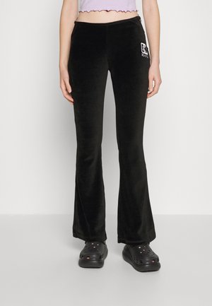 Black, flared velvet pants with a smooth texture and embroidered logo detail on the hip. Paired with black slip-on shoes.