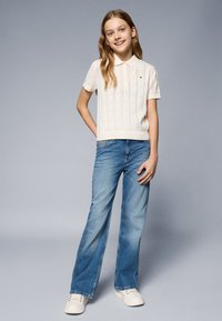 Polo de malha de manga curta, em creme, com padrão texturizado, combinado com jeans de perna larga azuis e ténis brancos.