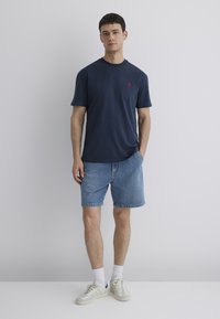 Dunkelblaue Baumwoll-T-Shirt mit kleinem rotem Logo, kombiniert mit hellblauen Jeanskurzshorts und weißen Socken, die lässige Ausstattung mit Turnschuhen vervollständigen.