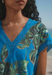 Haut bleu en v avec une bordure en dentelle, orné d'un motif de feuilles tropicales en vert et orange, confectionné en tissu léger.