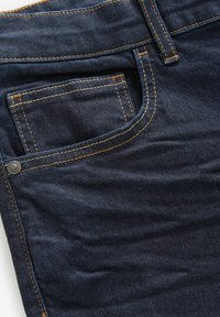 Jeans de mezclilla oscura con un bolsillo delantero, costuras amarillas en contraste y un acento de botón metálico en la cinturilla. Textura suave.