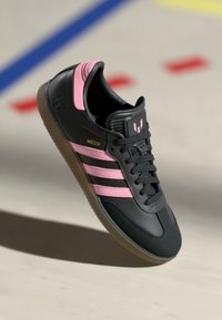 Sorte sneakers med pink accenter og tre striber. Har en gummisål og tekstureret hæl. "MESSI" logoet er vist på siden.