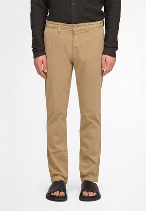 Mężczyzna ubrany w beżowe proste spodnie, czarną teksturowaną koszulę z długim rękawem i czarne damskie klapki z odkrytymi palcami, stojący na białym tle.