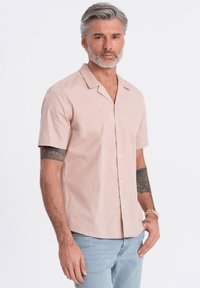 Korte mouwen, lichtroze shirt met een neklap en knopen aan de voorkant; gemaakt van een gladde stof. Draagt bij lichtblauwe jeans.