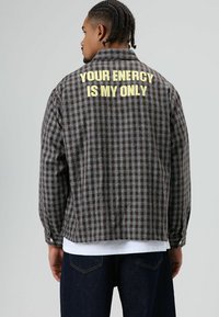 Bruin en grijs geruit flanellen jack met lange mouwen. Op de achterkant gele tekst: "YOUR ENERCY IS MY ONLY." Zachte textuur, ontspannen pasvorm.