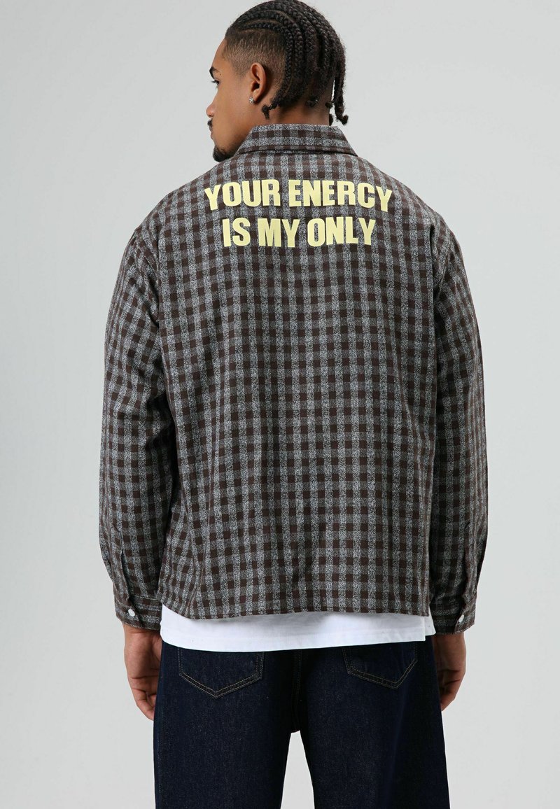 Bruin en grijs geruit flanellen jack met lange mouwen. Op de achterkant gele tekst: "YOUR ENERCY IS MY ONLY." Zachte textuur, ontspannen pasvorm.
