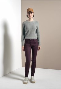 Pull en maille côtelée gris clair, pantalon slim-fit bordeaux et baskets beiges. Une casquette marron complète la tenue. Fond simple avec des teintes neutres.