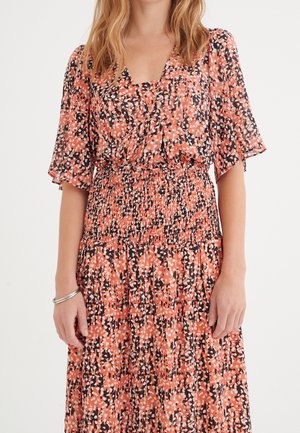 Kobieta ubrana w sukienkę z motywem koralowca, czarno-białą w cętki, z krótkimi, falistymi rękawami i marszczonym pasem, stojąca z luźno opuszczonymi rękami.