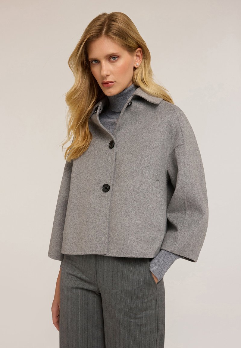 Giacca cropped in misto lana grigia con colletto, caratterizzata da due bottoni frontali e spalle cadenti, abbinata a pantaloni grigi a righe.