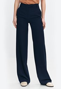 Granatowe spodnie w kant o szerokich nogawkach, wykonane z gładkiego materiału z eleganckim krojem, z średnim stanem. Połączone z beżowym obuwiem.