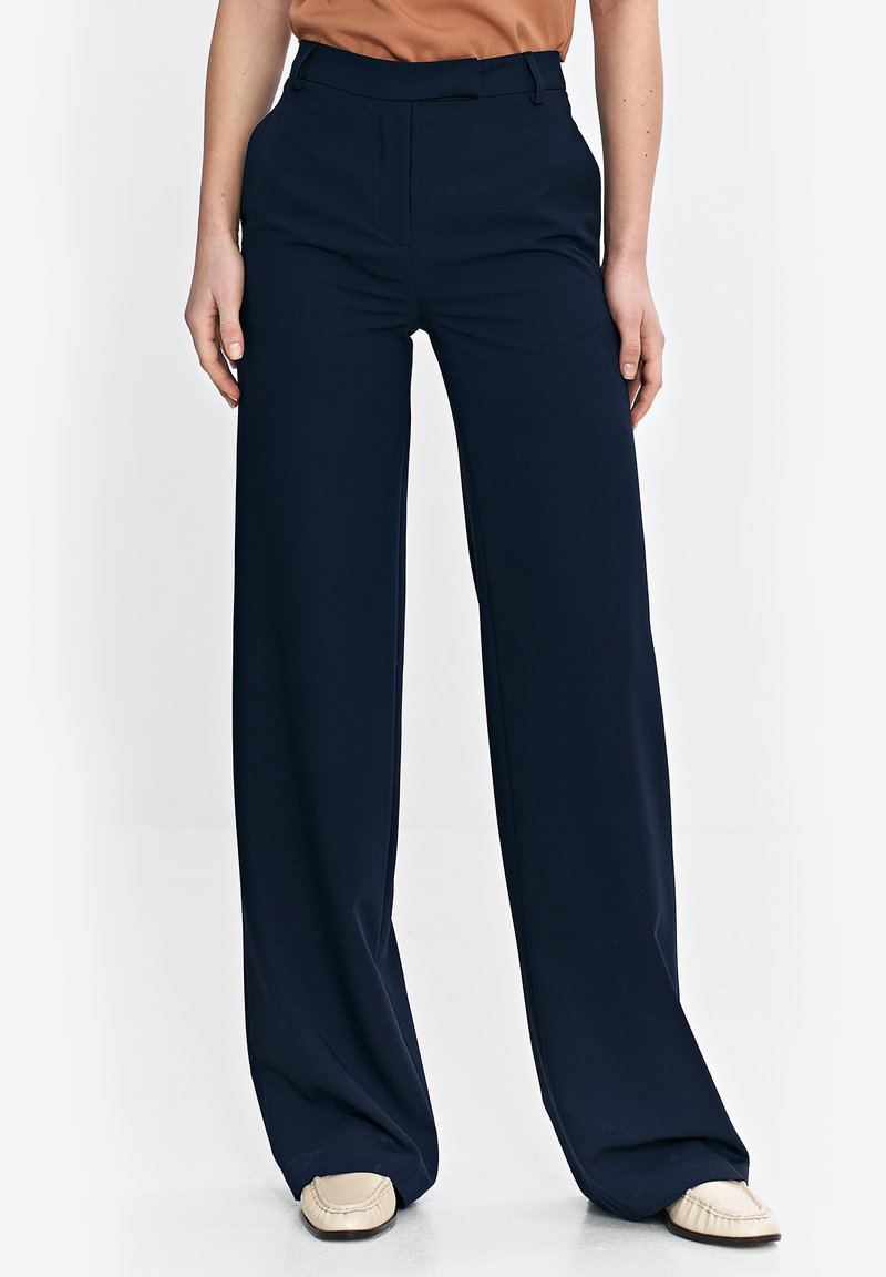 Granatowe spodnie w kant o szerokich nogawkach, wykonane z gładkiego materiału z eleganckim krojem, z średnim stanem. Połączone z beżowym obuwiem.