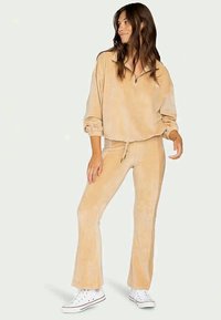 Haut zippé en velours côtelé beige avec des poignets élastiques, associé à un pantalon flare ajusté assorti. Le mannequin porte des baskets blanches.