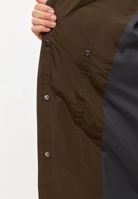 Tejido exterior marrón con una textura suave y forro interior gris. Presenta botones a presión y un diseño elegante en la sección abierta.
