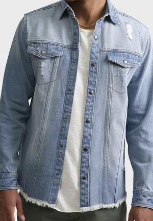 Homme portant une veste en jean bleu clair avec des bords effilochés et des détails usés sur une chemise blanche et un pantalon foncé.