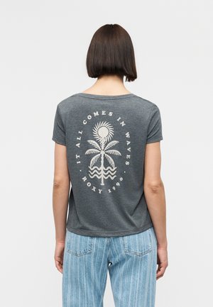 Kvinde med kort brunt hår iført en mørkegrå t-shirt med en hvid palme og sol-grafik samt teksten "Det hele kommer i bølger, Roxy 1990."