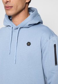 Lichtblauwe hoodie met een textuurafwerking, voorzien van een hexagonaal logo en een ritssluitingzak aan de linkermouw. Trekkoorden in bijpassende kleur.