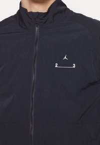 Gros plan d'une veste noire à fermeture éclair avec logo Jumpman blanc et « 2 3 » imprimé sur la poitrine, portée par une personne.