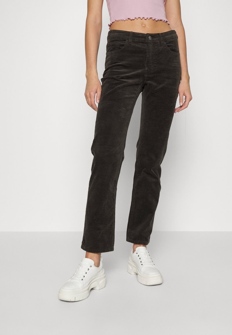 Pantalon en velours côtelé marron foncé avec une coupe droite, présentant des poches avant et une fermeture à bouton, associé à des baskets blanches.