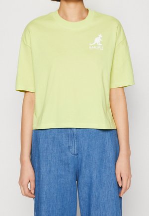 Osoba w jasnozielonym, krótkim, krótkim t-shircie Kangol z krótkimi rękawami i luźnych, wysokich, niebieskich spodniach, stojąca na jednolitym tle.