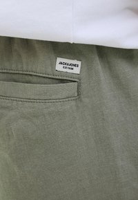 Pantalones de lino verdes con un bolsillo trasero que lleva una etiqueta que dice "JACK & JONES EST. 1990." Tejido texturizado con un acabado natural.