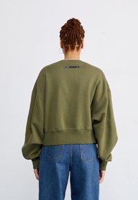 Maslinasto zelena oversized dukserica s spuštenim ramenima, rebrastim manžetama i rubom. Ima crnu oznaku na izrezu s logotipom brenda.