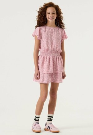 Jeune fille aux cheveux roux bouclés, portant une robe rose à motifs, des chaussettes blanches avec un logo, et des baskets roses, debout et souriante sur fond blanc.